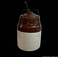 Front view of the stoneware crock showing two-tone brown and off-white glaze with attached metal handle and lid.