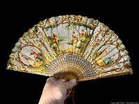 Front view of the opened antique Victorian paper lace folding fan held by a hand against a black background, showing detailed painted scenes, gold filigree, and the overall condition.