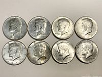 Obverse side of eight 1964 Kennedy half dollar coins showing the profile of President Kennedy and visible date. Coins appear circulated with some pink toning and wear.