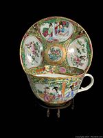 Front angled view of the teacup and saucer showing detailed hand-painted scenes and gold accents against dark background