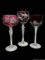 Three tall Bohemian glass goblets in red, purple, and pink with cut-to-clear floral and starburst designs.