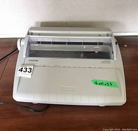 Top-view of Brother electronic typewriter with clear plastic cover and visible measurements on green tape.