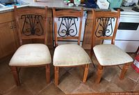 Front view of three wood framed dining chairs showing wrought iron decorative backs and cream upholstered seats with wear and rips.