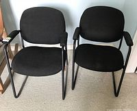 View of two black office chairs with metal frames side-by-side, showing fabric seats and backs with some wear and armrests