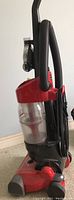 Side view of Dirt Devil vacuum showing red and black cyclonic upright design with clear dust canister and hose attached.