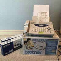 Front view of Brother IntelliFAX-775 plain paper fax machine with numeric keypad, function buttons, telephone handset attached, owner's manual inserted behind paper tray, original product box, and two ink cartridge boxes placed beside it.