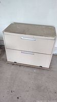 Front view of beige 2 drawer steel file cabinet sitting on wooden platform with casters, showing dusty top and clean-floor background.