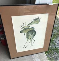 Framed print showing stylized moose artwork with brown mat and black frame. Glass and frame surface visible with some spots.