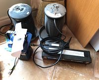 Two black cylindrical Kaz bug zappers, one black rectangular electric heater, and one small camping lantern together on floor.