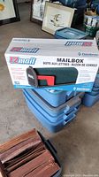 Photo of the boxed Flambeau EZmail mailbox on top of stacked blue plastic containers in a garage setting, showing the front and side packaging.