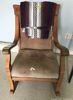 Wooden rocking chair with Mexican style throw draped over back, showing stained tan cushion seat and visible damage.