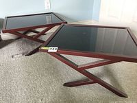 Two matching side tables with metal frames and black acrylic tops placed on carpeted floor, viewed at an angle showcasing the X-shaped leg design.