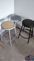 Set of three metal shop stools overall view, two with vinyl padded seats (black and gray), one with unpadded wooden seat (white frame).