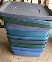 Stacked six Rubbermaid storage totes of varying blue, green and gray colors with lids. Shows dust and minor wear.
