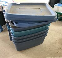 Stack of five plastic storage totes with lids in different shades of blue and teal, showing slight wear and dirt marks.