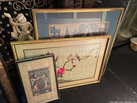 Three framed art pieces stacked against a wall with floral and architectural Asian art visible