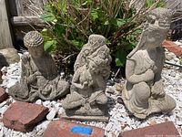 Three concrete Asian inspired statues displayed outdoors on white gravel with plants in the background. Shows all three statues in one view.