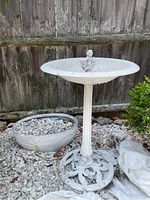 Wide view of white metal bird bath with ceramic planter pot beside it on gravel ground in front of wooden fence.