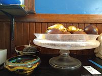 Pedestal frosted glass compote bowl holding wooden spheres and faux gold fruit, cloisonné bowl and small metal bowl with decorative stones in background