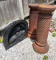 Plaster Buddha wall art with seated Buddha figure and two rust-colored twisted spiral decorative columns on gravel outdoors