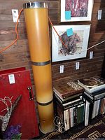 Full view of the tall cylindrical floor lamp in acrylic with gradient yellow-orange color, metal bands and cord, standing beside books and framed art.