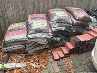Overall photo showing stacked bags of red rubber mulch along with a stack of red bricks on the ground beside them.