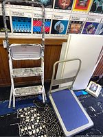 Photo showing a metal step ladder with white paint, four steps, heavily used, and metal hand truck with blue platform and beige frame.