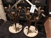 Front view of two metal cherub figures on white stone bases showing detailed wing and leg design