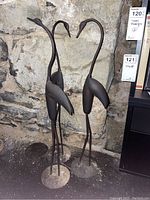 Three long-necked metal flamingo statues positioned against a stone wall showing two statues side by side.