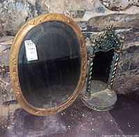 Photo showing both the oval wall mirror with a gold carved wooden frame and the small decorative tabletop mirror next to a stone wall.