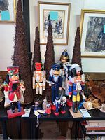 Photo showing multiple nutcracker figurines of different sizes and styles displayed on a black surface in front of brown cone-shaped decorations and framed abstract art.
