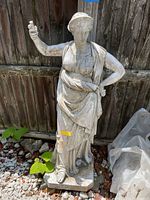 Full view of the outdoor ceramic statue standing against a wooden fence showing the overall figure and draped robes.