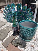 Photo showing metal peacock statue, ceramic planter, and stone reclining figure statue arranged outdoors on white gravel and bricks.