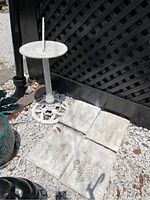 Overall view of white metal sundial on pedestal base next to four plaster tiles with fern imprints laid on ground with white gravel background.