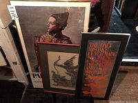 Full view of three framed art pieces leaning against a wall, showcasing diverse styles including poster, Asian art print, and mirrored quote.
