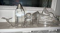 Five clear crystal and art glass figurines arranged on a white shelf: faceted prism, whale, bubble form, smaller prism, and running animal figure.