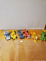 Wide shot of all six Hot Wheels cars in various colors and designs on wooden surface in front of cardboard background.