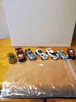 Seven different Hot Wheels cars arranged in a row on wooden surface with a white backdrop, showcasing variety and condition.