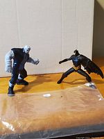 Two action figures on wooden surface: Batman black and yellow with wings, and pale grey muscular villain figure