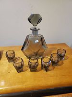 Full view of the smoky quartz decanter with stopper and six matching faceted glasses arranged around it on a wooden surface.