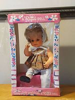 Vintage Baby Jill doll posed inside original pink and floral cardboard display box showing the doll's outfit and seated position.