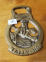 Front view of vintage brass horse brass featuring a rider on horse jumping a fence and 'TALLY-HO' inscription