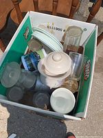 Box containing various drinking glasses, a ceramic bowl, a ceramic jar with lid, and a printed ceramic mug.