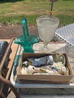Green glass basket, clear diamond-cut goblet, porcelain figurines in box and glass square item shown on table.