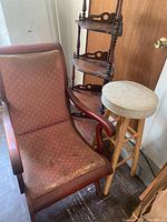 Photo showing a wooden armchair with reddish fabric upholstery next to a wooden stool with cushioned seat, and a three-tier wooden corner shelf.