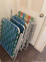 Photo of six vintage lawn chairs folded and stacked against a white door, showing woven plastic webbing in green, blue, orange, and yellow colors with aluminum frames.