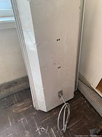 Two large rectangular white metal overhead garage light fixtures shown standing vertically against wall with attached power cords visible, placed in a corner with nearby window and patterned floor tiles.