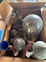 View inside one cardboard box showing a vintage glass dome clock with visible brass mechanism, a ceramic stein with metal lid, and vintage oil lamps with glass chimneys alongside other metal and glass items.