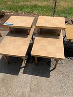 Four wooden side tables with square checkered tops and tapered cylindrical legs arranged on a concrete surface outside with grass in the background.