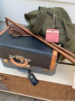 Full view of vintage black suitcase with green army duffle bag, leather belts, and wooden walking canes on top